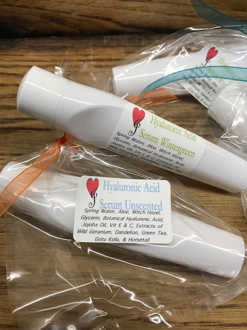 May include: Two white tubes of serum with labels that say "Hyaluronic Acid Serum Wintergreen" and "Hyaluronic Acid Serum Unscented". The labels have a red heart with a musical note in it. The tubes are wrapped in clear plastic with orange and teal ribbon.