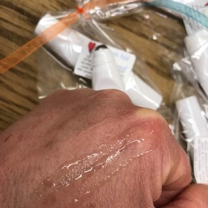 May include: A clear, colorless lip balm tube with a white cap. The balm has been applied to a hand, leaving a clear, shiny streak.
