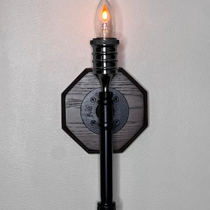 Może przedstawiać: Lampa ścienna z czarną metalową ramą i dekoracyjnym drewnianym oparciem. Oprawa posiada przezroczystą żarówkę w kształcie płomienia, która emituje ciepłą, pomarańczową poświatę. Oprawa została zaprojektowana w stylu vintage i industrialnym.