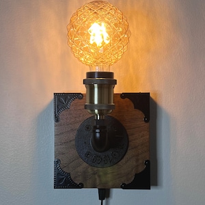 Puede incluir: Un aplique de pared de estilo vintage con una bombilla decorativa ámbar texturizada. La bombilla está colocada en un casquillo de latón, montada sobre una base cuadrada de madera con escuadras metálicas y un racor de tubería negro. Un cable negro sale de la base.