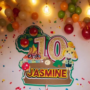 May include: A colorful birthday cake topper featuring the number 10 in gold glitter, the name "Jasmine", and cartoon characters. The topper is decorated with red apples and green leaves. Balloons and confetti are in the background.
