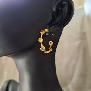 May include: Gold hoop earrings with colorful gemstones. The earrings have a circular shape and are adorned with small, round stones in shades of red, blue, green, and white.