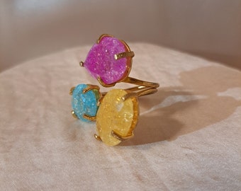 May include: Three gold rings with colourful crystal stones. The rings are stacked on top of each other. The top ring has a pink stone, the middle ring has a blue stone, and the bottom ring has a yellow stone.