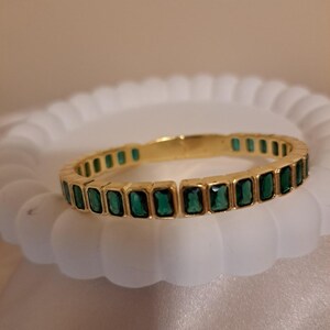 Könnte beinhalten: Ein goldenes Armreif-Armband mit einer Reihe rechteckiger smaragdgrüner Steine.