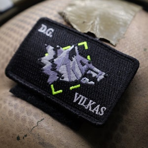 May include: A black patch with a stylized wolf design in purple and a green border. The patch has the text "VILKAS" and "DG" in white.