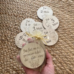 May include: A set of wooden baby milestone discs, each with engraved numbers and sea creature designs. The set includes discs for months 1-9, plus a disc with French text. The discs are packaged in a sheer bag.