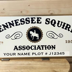 May include: A wooden sign in the shape of the state of Tennessee. The sign is white with black text that reads "Tennessee Squire Association" and includes a logo of a person riding a horse. The sign also includes the text "Your Name Plot # J12345".