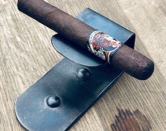May include: A metal cigar holder with a dark gray finish. The holder has a curved base and a raised lip to hold the cigar. A brown cigar is resting in the holder.