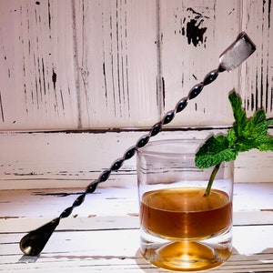 May include: A black metal cocktail stirring spoon with a twisted handle and a flat, rounded end. The spoon is resting on a white surface next to a glass of brown liquid with a sprig of fresh mint.
