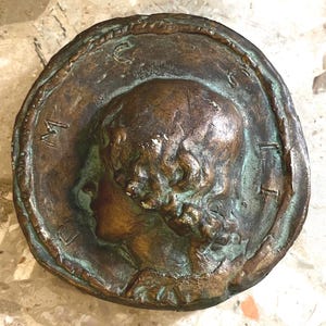 May include: A circular bronze-toned medallion featuring a relief portrait of a person in profile. The surface has a weathered, oxidized patina with green and brown tones. The letters M, C, and I are embossed along the textured, raised border.