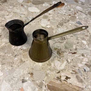 May include: Two small, vintage coffee pots with long handles. One is black, the other is brass-colored. The pots are sitting on a speckled, light-colored surface. The handles have a small hole at the end.
