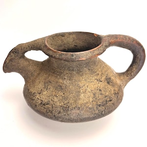 May include: A small, round, earthenware vessel with a textured, mottled brown and tan surface. It features a wide, open circular mouth, a short spout on the left side, and a curved handle on the right side, creating a symmetrical, rustic appearance.