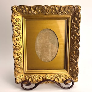 May include: A gold-colored, ornate picture frame with an oval opening. The frame has detailed scrollwork and floral accents. It is displayed on a dark brown metal stand, suitable for a photograph or artwork.