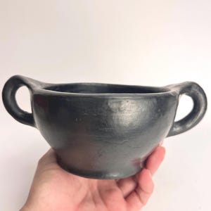 May include: A small, matte black ceramic bowl featuring two rounded handles on opposite sides. The bowl has a smooth, dark finish with subtle variations in texture across the surface, a wide circular opening, and a slightly tapered base.