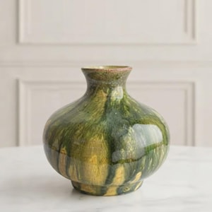 May include: A ceramic vase with a rounded body and a short, narrow neck. The surface features a glossy, marbled glaze with blended streaks of deep forest green, olive, and golden yellow tones, creating a fluid, organic appearance.