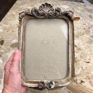 May include: Ornate rectangular picture frame with a distressed silver and gold finish. The frame features a shell-shaped design at the top and scrollwork details. The frame is empty, ready to display a photograph or artwork.