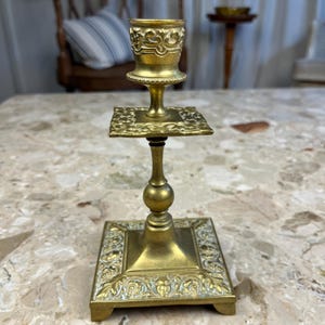 Könnte beinhalten: Ein Vintage-Kerzenhalter aus Messing mit quadratischer Basis und dekorativem floralem Design. Der Kerzenhalter hat einen hohen, schlanken Stiel mit rundem Mittelteil und einer kleinen Tasse oben. Die Gesamthöhe beträgt ca. 18 cm.