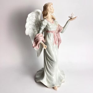 May include: A porcelain figurine of an angel with long blonde hair, wearing a flowing pale blue gown with a pink sash and accents. The angel has large white wings and holds a small white dove in one outstretched hand, standing in a pose.