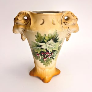 May include: A cream-colored ceramic vase featuring two sculpted ram heads on the sides. The body is decorated with a central floral motif of green leaves and small pink flowers. The rim and base have a warm, golden-yellow gradient finish.