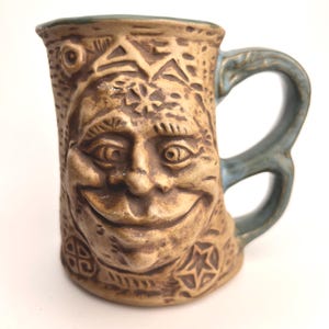 May include: A tan ceramic mug with a smiling face carved into the front. The mug has a blue handle and rim, and the face is surrounded by carved geometric patterns. The mug is a unique piece of pottery.