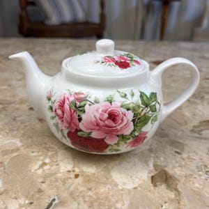 Könnte beinhalten: Weiße Keramik-Teekanne mit Deckel, verziert mit einem floralen Rosenmuster in Rosa und Rot, akzentuiert mit grünen Blättern. Die Teekanne hat einen gebogenen Griff und Ausguss, ideal zum Servieren von Tee.