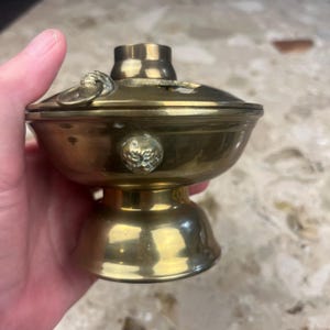 May include: A brass incense burner with a rounded bowl, a pedestal base, and a removable lid featuring a central chimney-style opening. The exterior of the bowl is decorated with small, raised circular motifs and a metal ring handle on the lid.