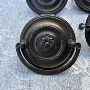 以下が含まれることがあります： 中央に花の模様、周囲にロープのような縁取りが施された、アンティークブロンズの引き出し取っ手。取っ手はカーブしたハンドルで、キャビネットやドレッサー用と思われる。この金具はヴィンテージ感がある。