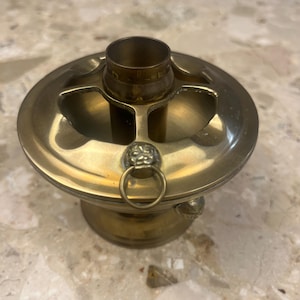 May include: A vintage brass ashtray with a circular design. It features a central cylindrical holder and three curved rests. The ashtray has a decorative lion's head with a ring handle and is elevated on a pedestal base.