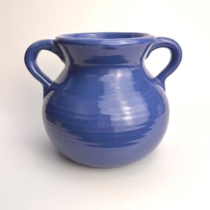 Könnte beinhalten: Eine blaue Keramikvase mit zwei Henkeln. Die Vase hat einen abgerundeten Körper und eine weite Öffnung. Die Außenseite hat eine glänzende Oberfläche und eine strukturierte Oberfläche. Die Henkel sind gebogen und am oberen Teil der Vase befestigt. Die Vase eignet sich für die Inneneinrichtung.