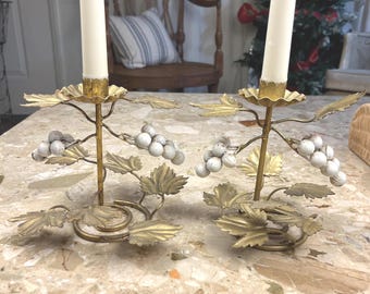 Puede incluir: Un par de candelabros de metal dorado con racimos de uvas y hojas de parra esculpidas. Cada candelabro tiene un tallo central que sostiene una vela blanca cónica, con una base circular y detalles de hojas que rodean las uvas.