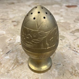 May include: A vintage brass egg-shaped salt shaker with a decorative floral pattern and small holes on top. The shaker has a pedestal base and is displayed on a marble surface. The brass has a warm, aged patina.
