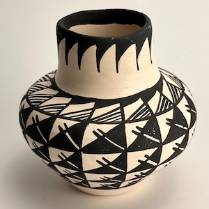 May include: A ceramic vase with a rounded body and a narrow neck, featuring a matte off-white base colour. The surface is decorated with hand-painted black geometric patterns, including triangular shapes and fine line details around the neck.