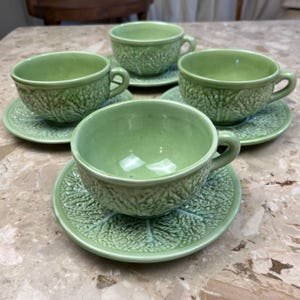 Puede incluir: Cuatro tazas de té y platillos de cerámica verde claro. Las tazas y los platillos tienen un diseño texturizado de hoja de col. Las tazas tienen un asa y se asientan sobre platillos a juego. El juego está sobre una superficie similar al mármol.