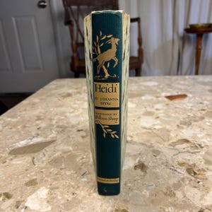 May include: A vintage hardcover book titled "Heidi" by Johanna Spyri, illustrated by William Sharp. The spine is dark green with gold lettering and a gold illustration of a deer. The book's cover has a cream-colored pattern.