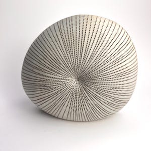 May include: A white, rounded ceramic sculpture with a textured surface. The surface is covered in a pattern of black, dashed lines radiating from a central point. The sculpture has a smooth, matte finish.