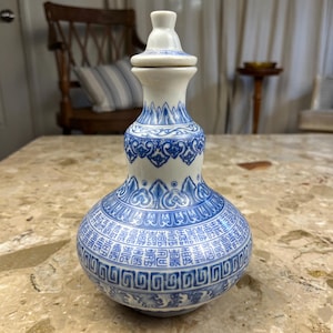 May include: A white and blue ceramic vase with a lid. The vase has a bulbous body and a long neck. The design features blue patterns and Chinese characters. The vase is approximately 10 inches tall.