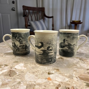 May include: Three ceramic mugs with wildlife illustrations. One mug features a deer, another depicts ducks, and the third shows ducks in a pond. The mugs have a cream-colored rim and handle, with black and white imagery.