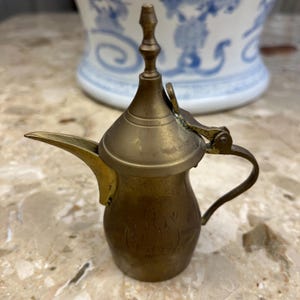 May include: A small, antique brass coffee pot with a curved spout, handle, and hinged lid. The pot has etched detailing and a decorative finial on top. The background features a white and blue patterned ceramic piece.