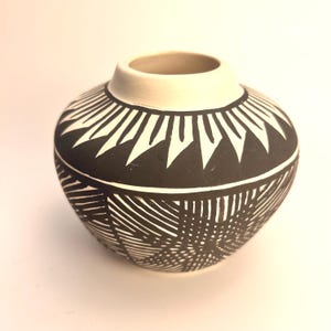 May include: A small, rounded ceramic vase featuring a matte black finish with intricate white geometric patterns. The upper section displays a series of sharp, triangular shapes, while the lower body is covered in fine, intersecting linear designs.