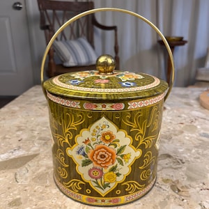 Peut inclure: Une boîte en métal vert olive vintage avec une poignée et un couvercle dorés. La boîte présente des motifs floraux orange, rose, jaune et bleu. La poignée et le bouton du couvercle sont dorés. La boîte est probablement utilisée pour le stockage ou l'affichage.