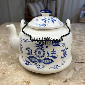 May include: A white ceramic teapot with blue floral patterns and a black wire handle. The lid is topped with a blue knob. The teapot has a spout and is decorated with blue floral designs.