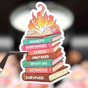 May include: A die-cut sticker featuring a stack of seven books with flames rising from the top book. The book spines are labeled with the words Anxiety, Overthinking, Stress, Mistakes, What-ifs, Setbacks, and Survived in white capital letters.