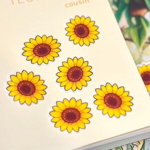 May include: A cream-colored planner cover with the text "HOBO TECHO 2024 cousin" printed in light brown. Six individual sunflower stickers with bright yellow petals and dark brown centers are arranged across the surface of the planner.