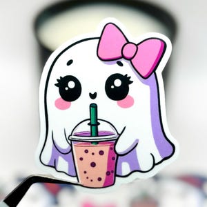 May include: A white ghost-shaped sticker with a pink bow, large eyes, and rosy cheeks. The ghost is drinking a pink bubble tea with a green straw. The sticker is held by a pair of tweezers, with other stickers in the background.