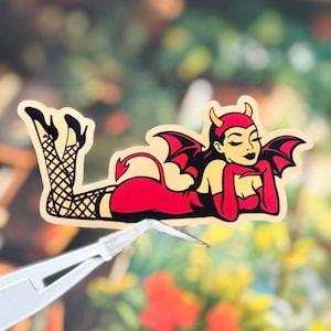 May include: A die-cut sticker featuring a cartoon devil character with red skin, black bat wings, and a pointed tail. The character wears a red dress, black high heels, and black fishnet stockings while lying down in a reclining pose with eyes closed.