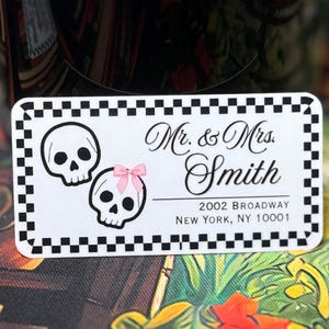 May include: A rectangular address label with a black and white checkered border. It features two white skulls, one with a small pink bow, and the text: Mr. & Mrs. Smith, 2002 Broadway, New York, NY 10001, printed in a black script and sans-serif font.