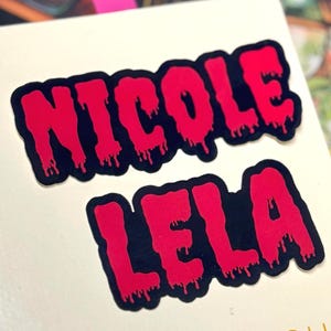 May include: Two stickers featuring the names NICOLE and LELA in a bright pink, dripping font style with a thick black outline. The letters have a liquid, melting appearance, creating a bold graphic effect against the light-colored surface.