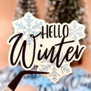 May include: A die-cut sticker featuring the text "HELLO Winter" in a dark brown script and sans-serif font. The background is cream-colored and decorated with light blue snowflake illustrations. The sticker is held by a pair of metal tweezers.