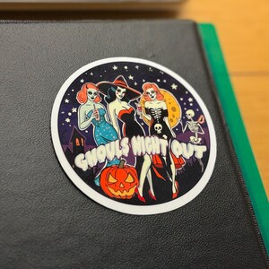 May include: A circular sticker featuring a Halloween-themed illustration of three stylized figures in costumes against a dark starry night sky with a full moon, a jack-o'-lantern, and a skeleton, with the text GHOULS NIGHT OUT printed in white.
