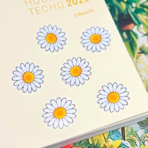 May include: Six daisy stickers with white petals and yellow centers arranged on a cream-colored planner cover. The planner features the text "HOBO TECHO 2023 cousin" printed in light brown at the top. The stickers are circular and flat.